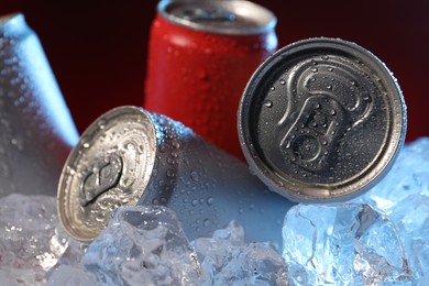Photo of Refreshing drinks in cans on ice cubes against dark red background, closeup