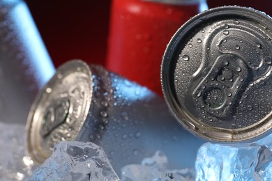 Photo of Refreshing drinks in cans on ice cubes against dark red background, closeup