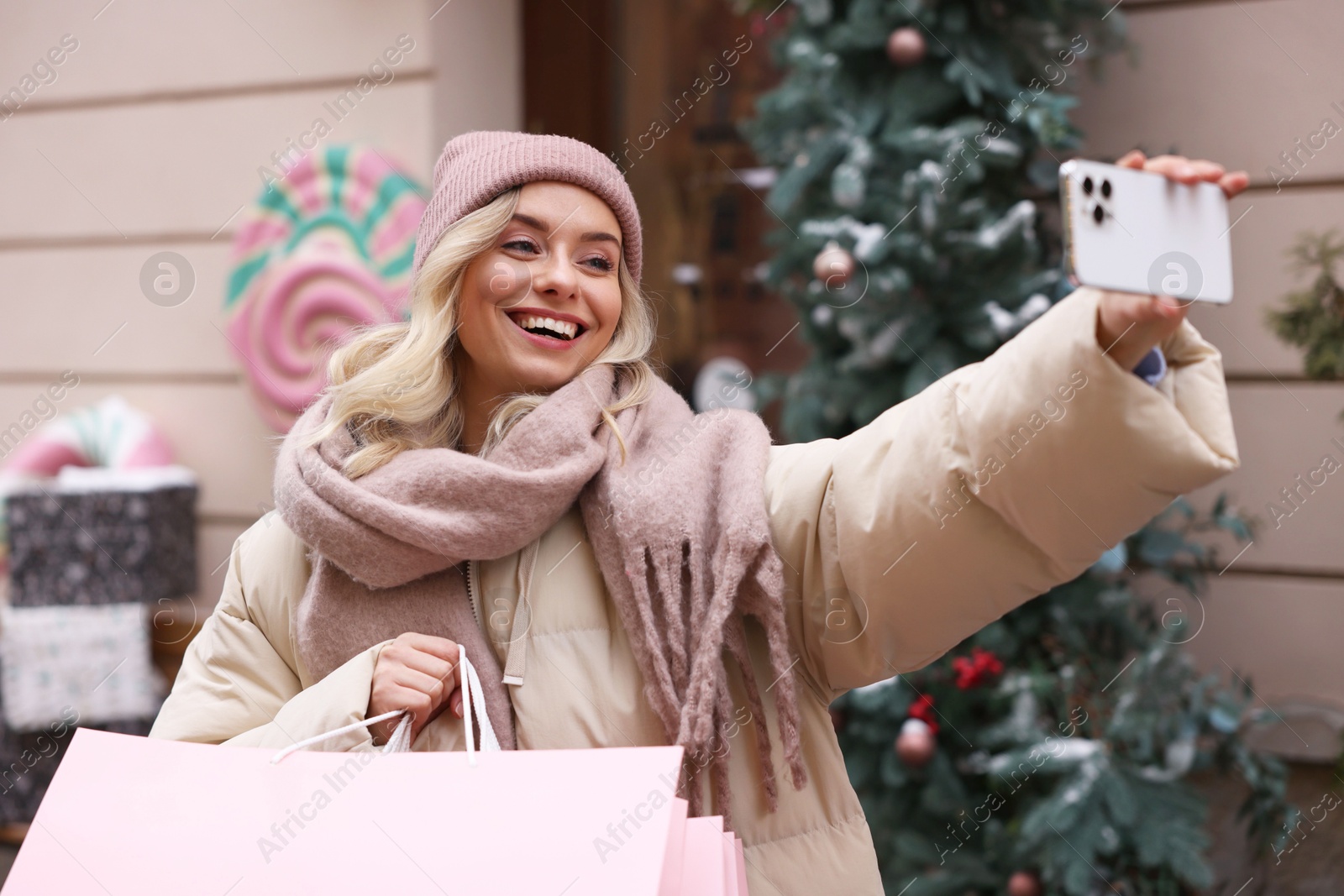 Photo of Happy woman with paper bags taking selfie indoors. Christmas season