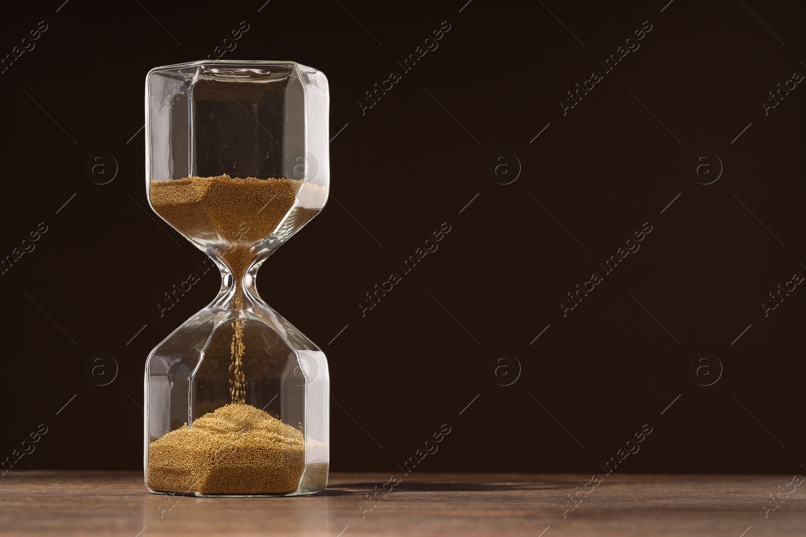 Photo of Hourglass with flowing sand on wooden table against dark brown background, space for text