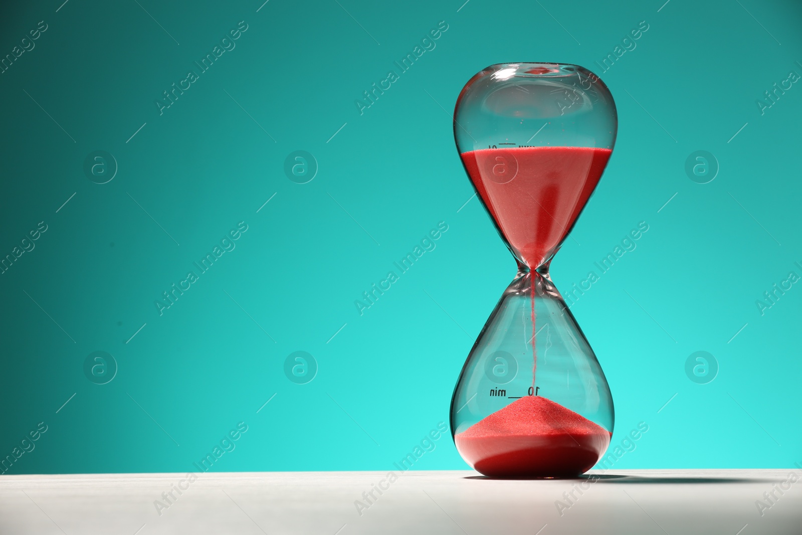 Photo of Hourglass with red flowing sand on white table against turquoise background. Space for text