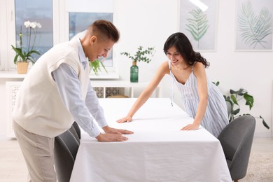 Photo of Couple putting white tablecloth on table at home