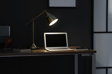 Photo of Comfortable workplace with laptop and lamp on wooden desk at home office in evening