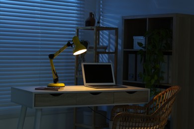 Photo of Comfortable workplace with laptop and lamp on desk at home office in evening