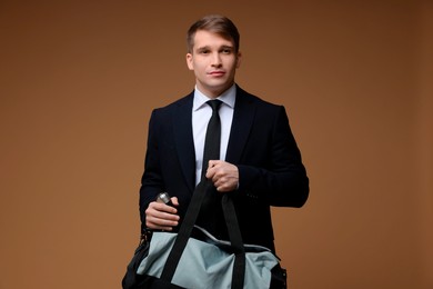 Young man in suit putting bottle of water into gym bag on brown background