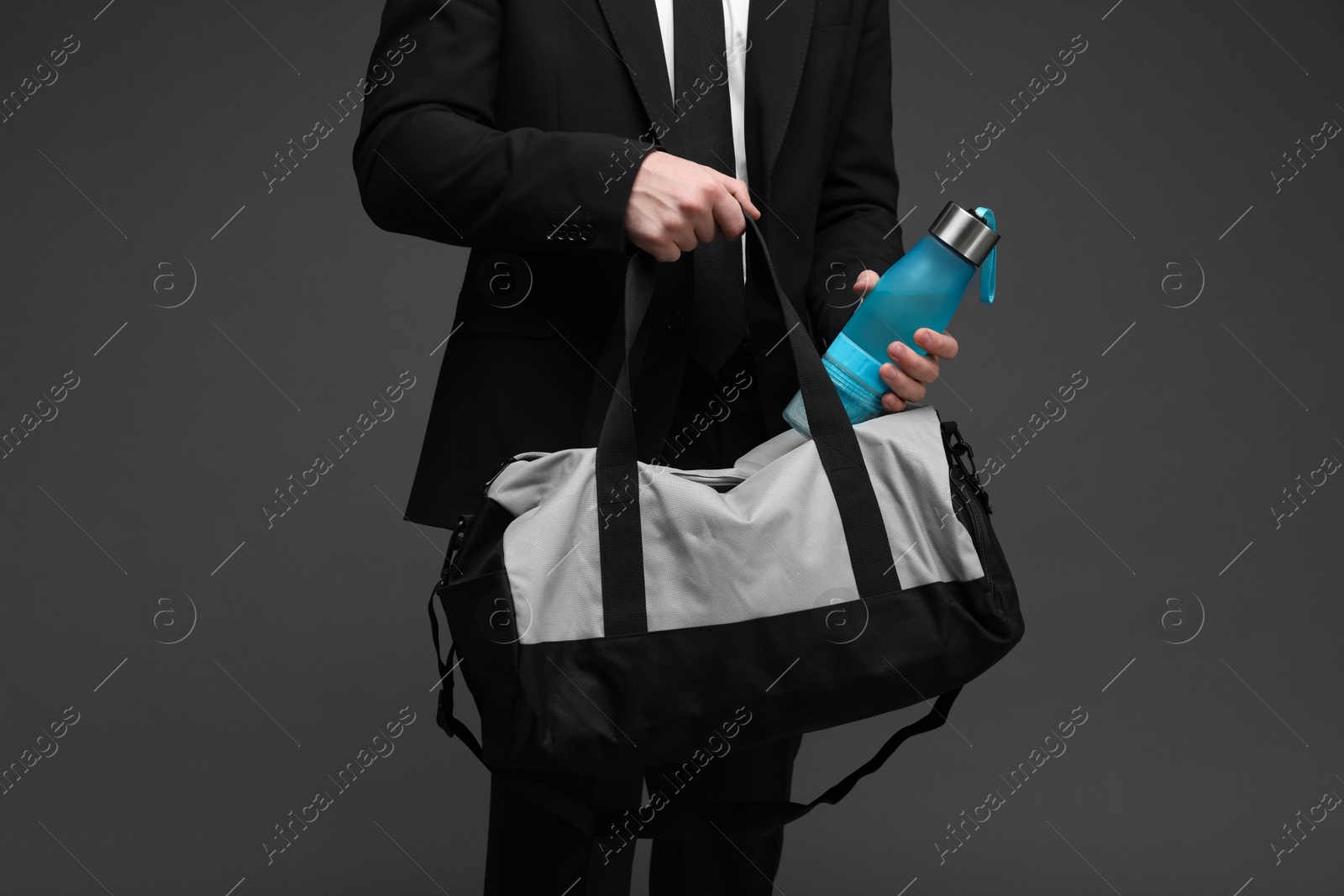 Photo of Man in suit putting bottle of water into gym bag on dark grey background, closeup