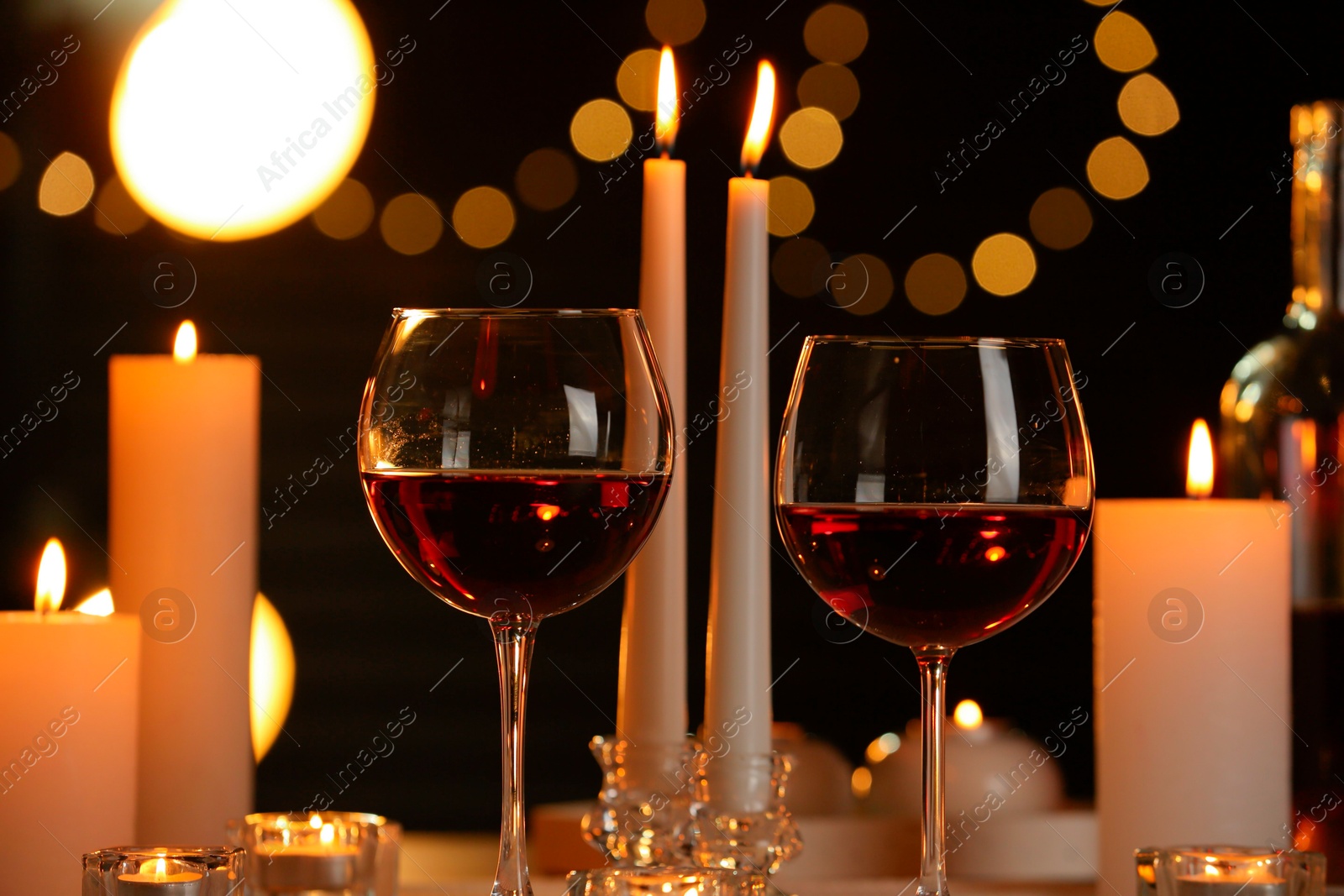 Photo of Glasses of red wine and burning candles against blurred lights, closeup. Romantic dinner