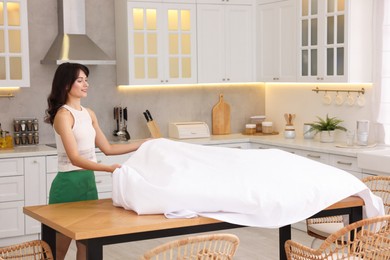 Photo of Young woman putting white tablecloth on table in kitchen