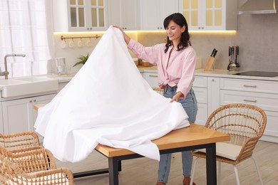 Photo of Young woman putting white tablecloth on table in kitchen