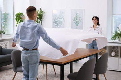 Couple putting white tablecloth on table at home