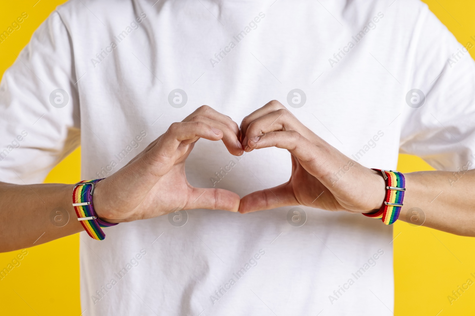 Photo of Man with bracelets in colors of LGBT flag making heart gesture on yellow background, closeup