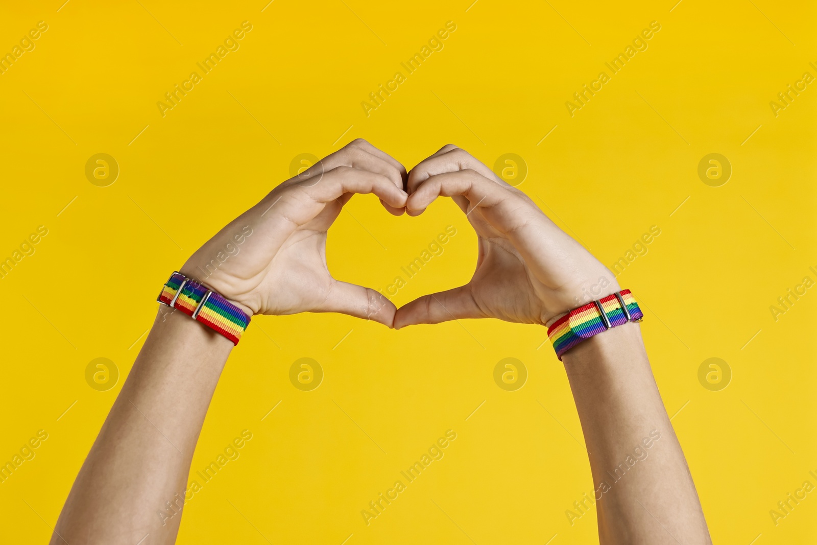 Photo of Man with bracelets in colors of LGBT flag making heart gesture on yellow background, closeup