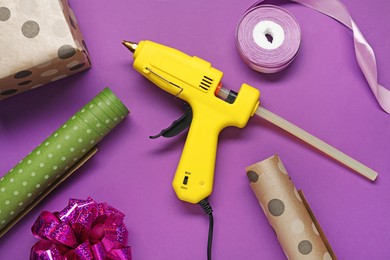 Photo of Hot glue gun and handicraft materials on purple background, flat lay