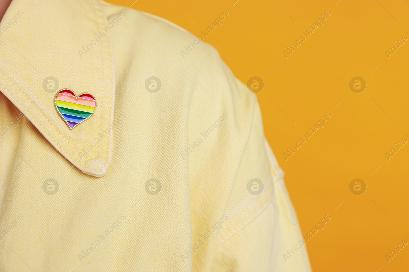 Photo of Woman with heart shaped pin in LGBT colors on orange background, closeup. Space for text