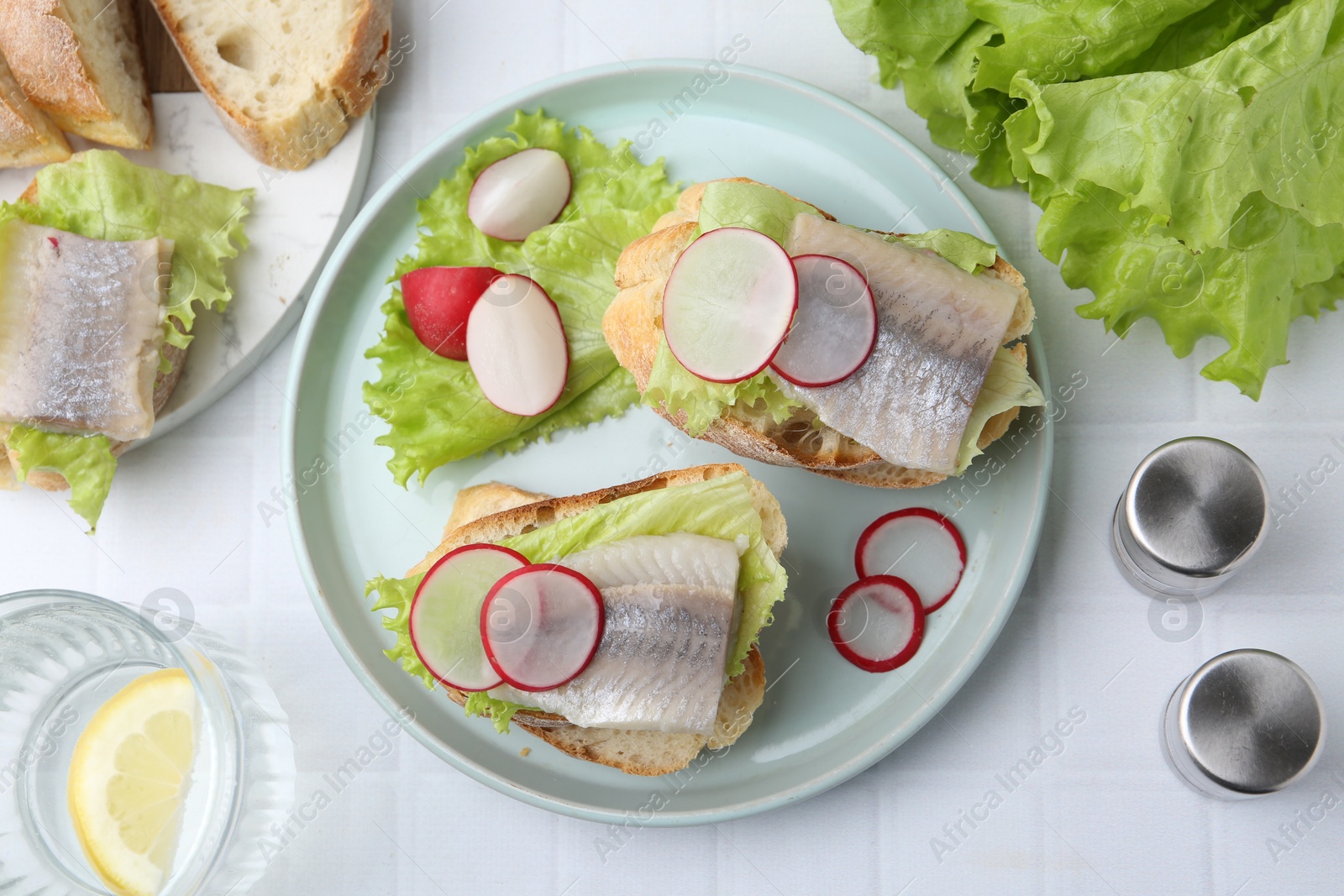 Photo of Tasty sandwiches with herring, radish and lettuce on white tiled table, flat lay