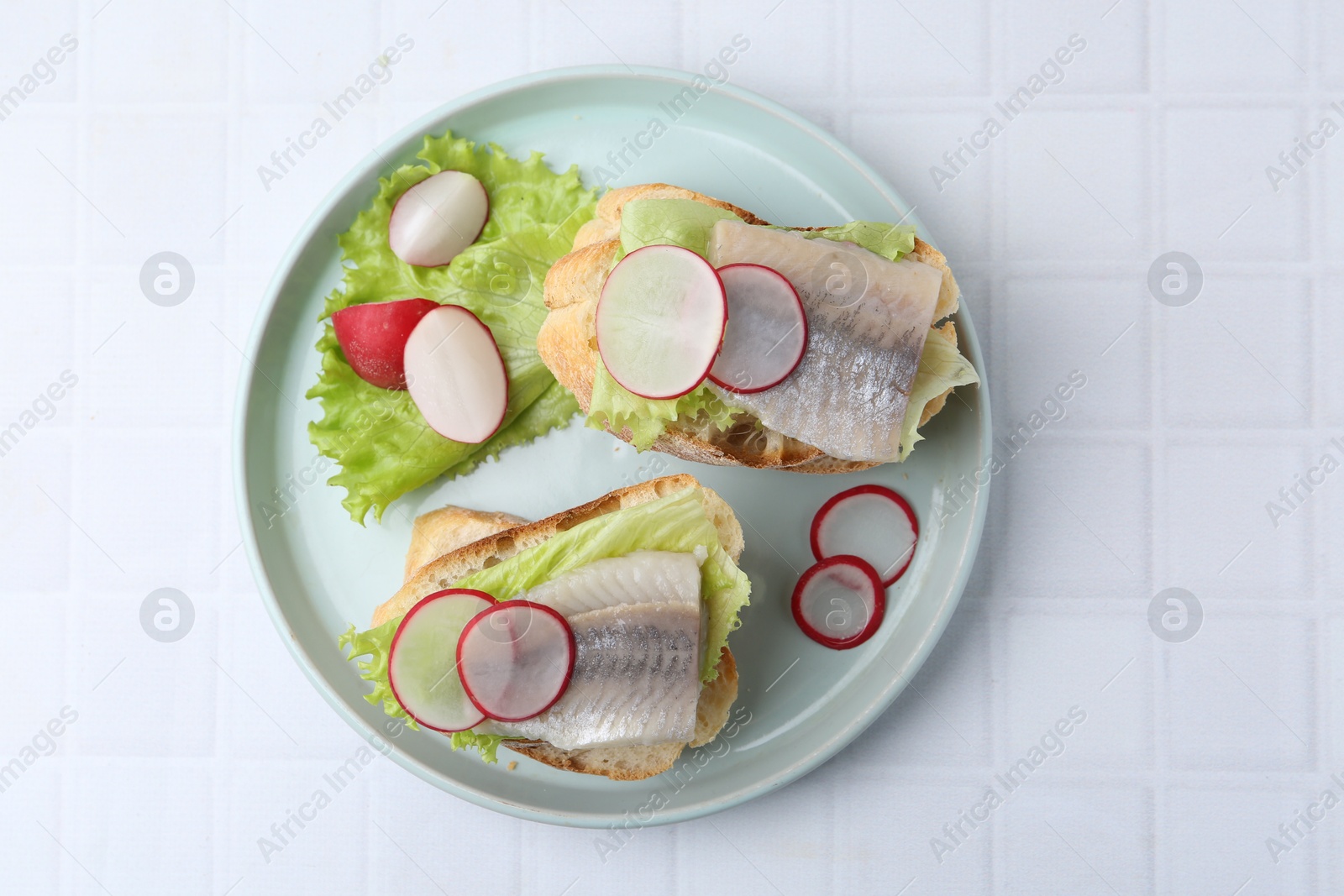 Photo of Tasty sandwiches with herring, radish and lettuce on white tiled table, top view