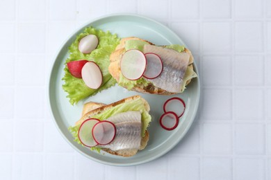 Photo of Tasty sandwiches with herring, radish and lettuce on white tiled table, top view