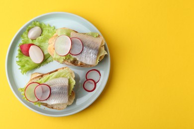 Photo of Tasty sandwiches with herring, radish and lettuce on yellow background, top view. Space for text