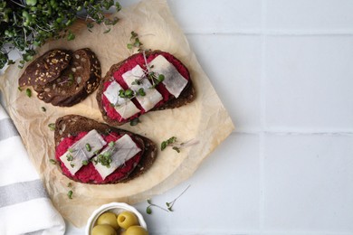 Photo of Tasty sandwiches with herring, horseradish sauce and olives on white tiled table, flat lay. Space for text