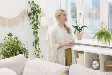 Photo of Feng shui. Young woman with glass of water near houseplants at home