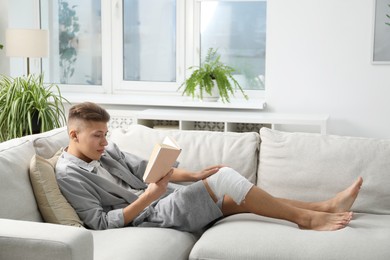 Photo of Man with medical bandage on his knee reading at home