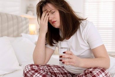 Unhappy woman with glass of water suffering from hangover on bed at home