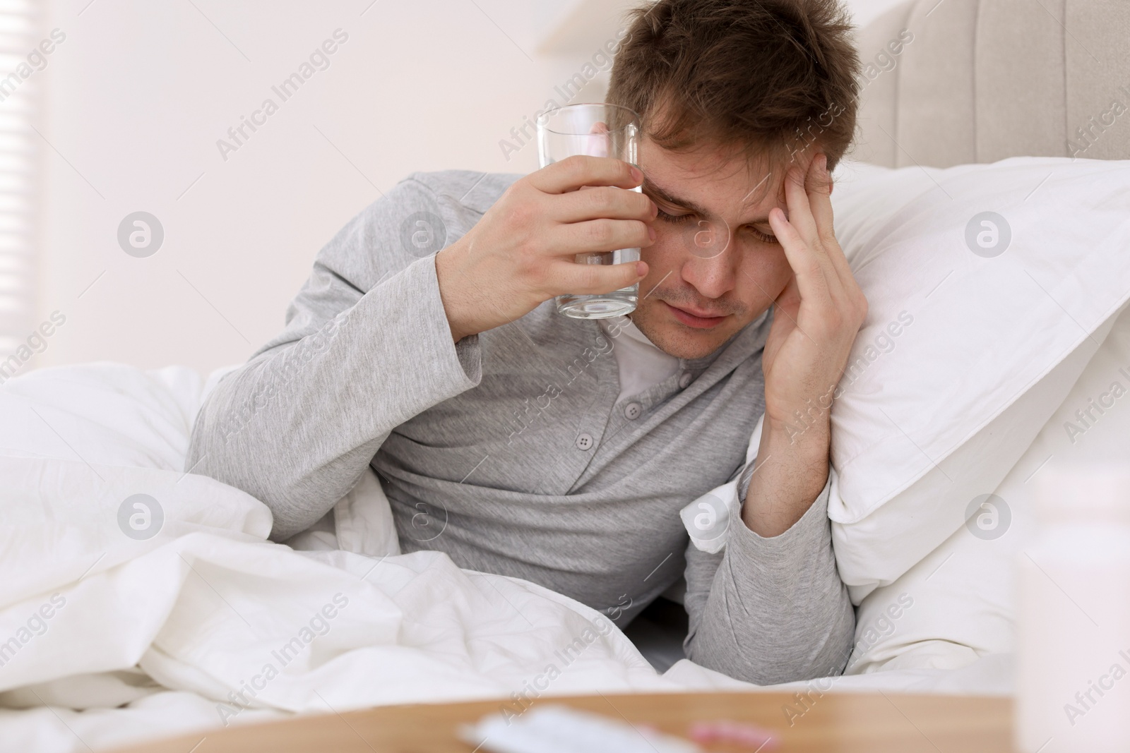 Photo of Unhappy young man with glass of water suffering from hangover on bed at home