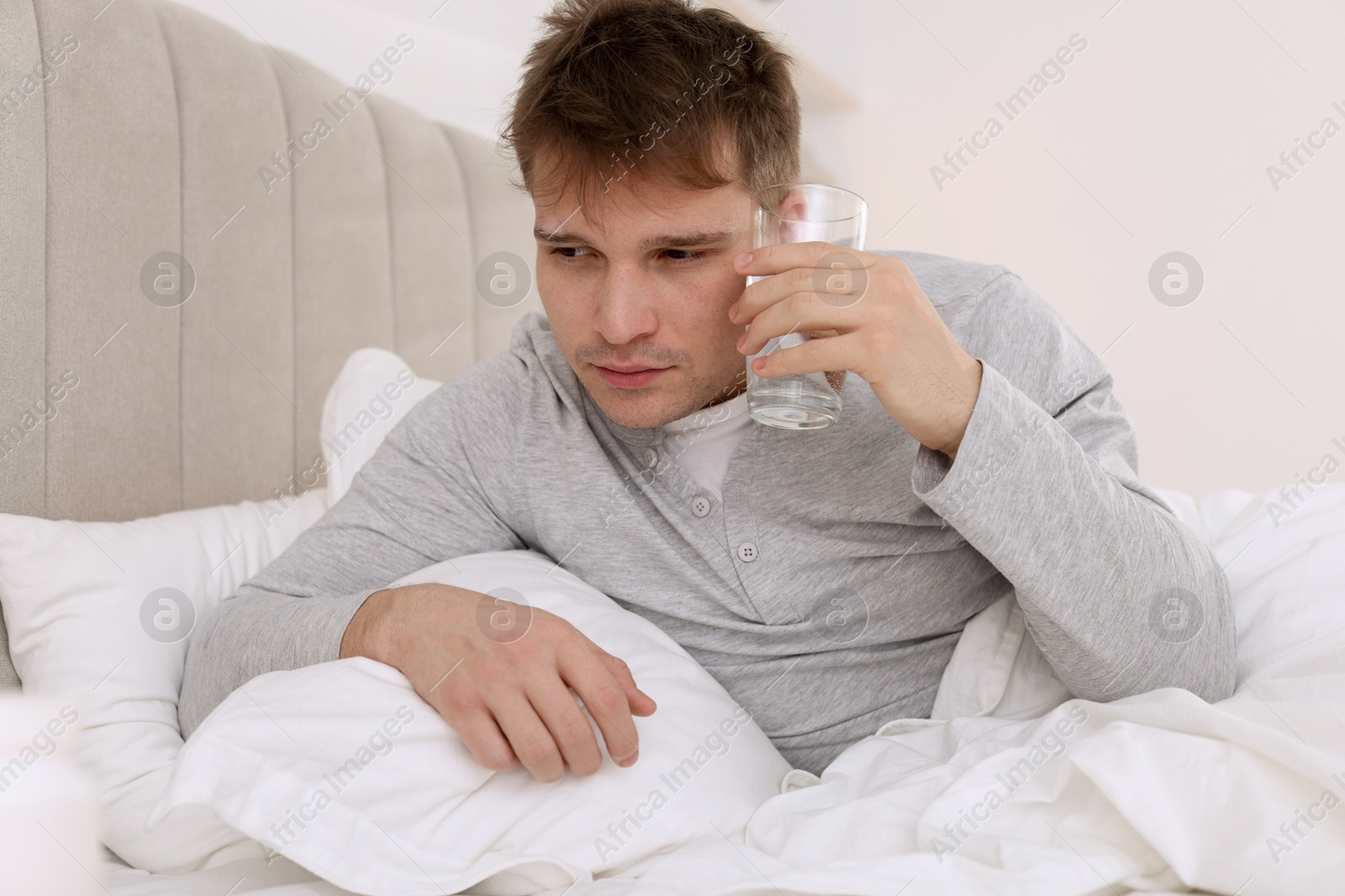 Photo of Unhappy young man with glass of water suffering from hangover on bed at home