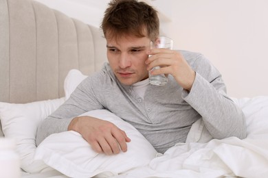 Photo of Unhappy young man with glass of water suffering from hangover on bed at home