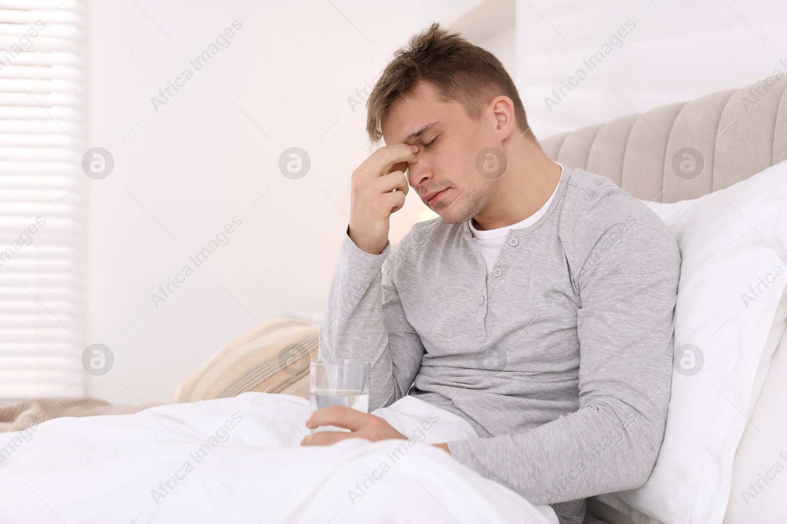 Photo of Unhappy young man with glass of water suffering from hangover on bed at home, space for text