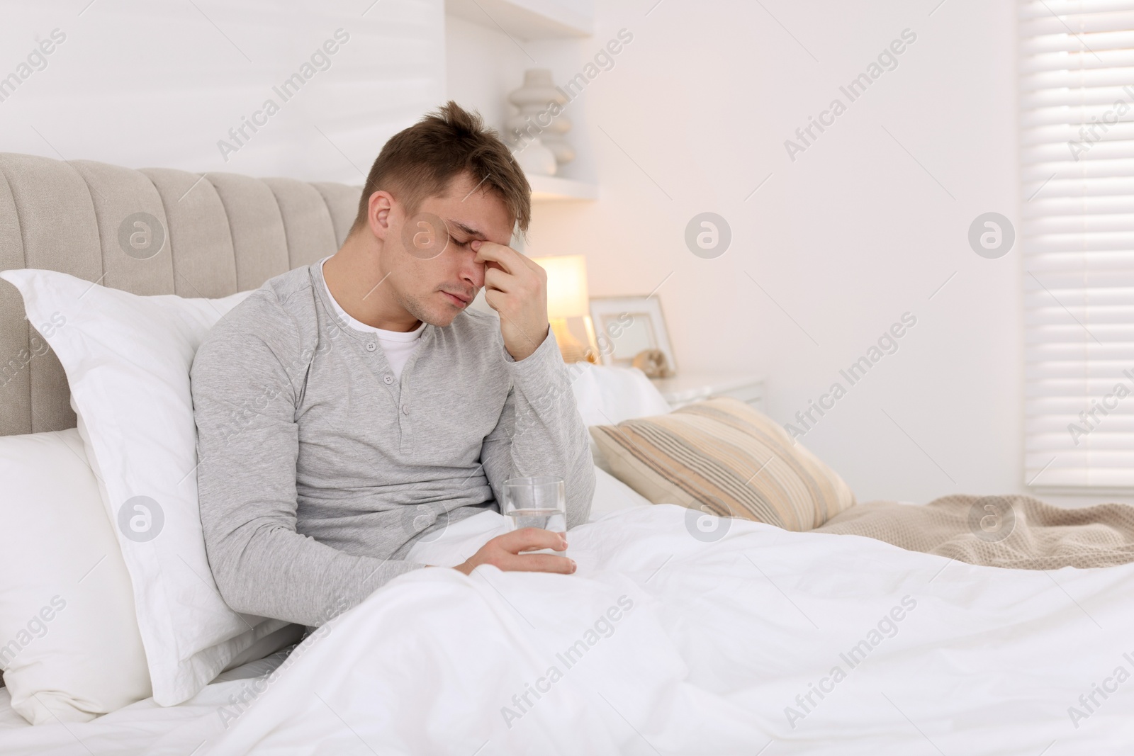 Photo of Unhappy young man with glass of water suffering from hangover on bed at home, space for text