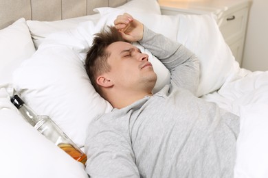 Photo of Unhappy young man with bottle of whiskey suffering from hangover on bed at home