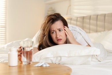 Unhappy woman with glass of water and pills suffering from hangover on bed at home