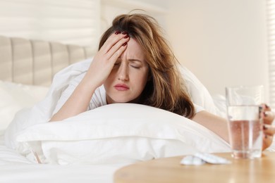 Photo of Unhappy woman with glass of water and pills suffering from hangover on bed at home