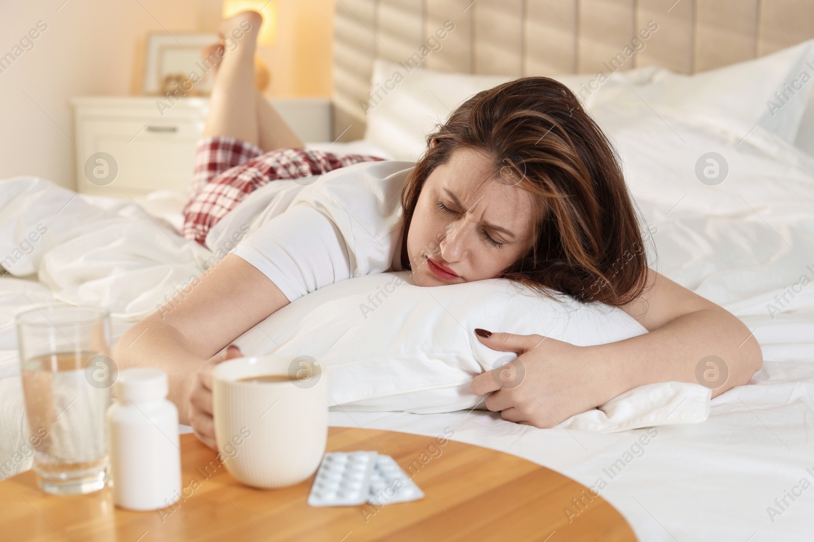 Photo of Unhappy woman with cup of coffee, water and pills suffering from hangover on bed at home