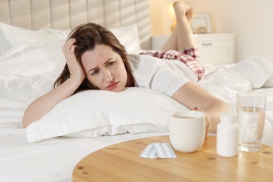 Photo of Unhappy woman with cup of coffee, water and pills suffering from hangover on bed at home