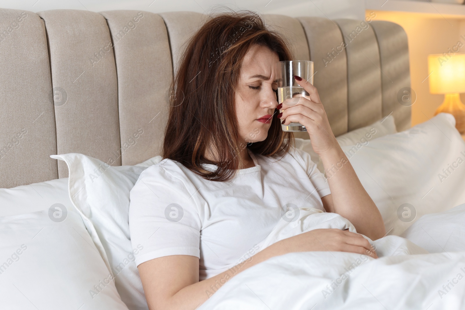 Photo of Unhappy woman with glass of water suffering from hangover on bed at home