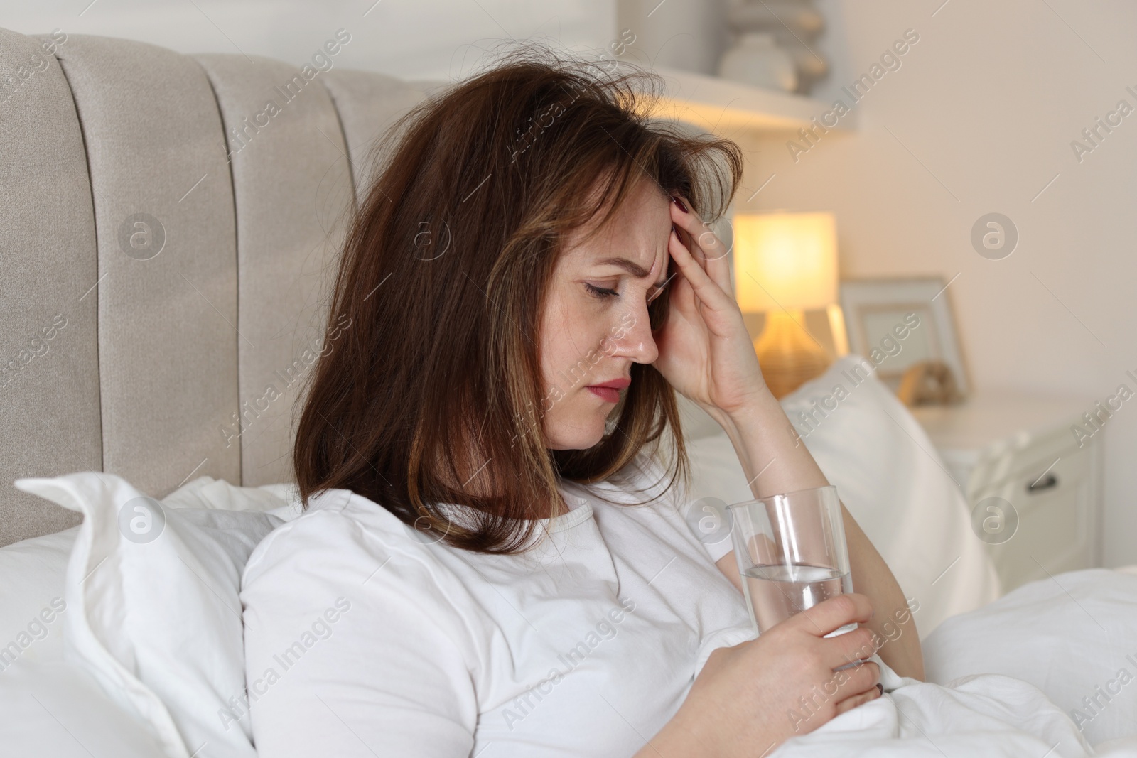 Photo of Unhappy woman with glass of water suffering from hangover on bed at home