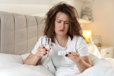 Photo of Unhappy woman with pills and glass of water suffering from hangover on bed at home