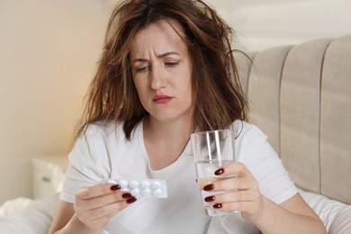 Photo of Unhappy woman with pills and glass of water suffering from hangover on bed at home