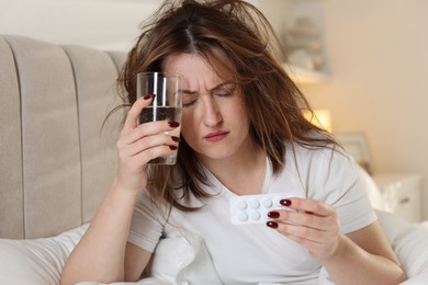 Photo of Unhappy woman with pills and glass of water suffering from hangover on bed at home