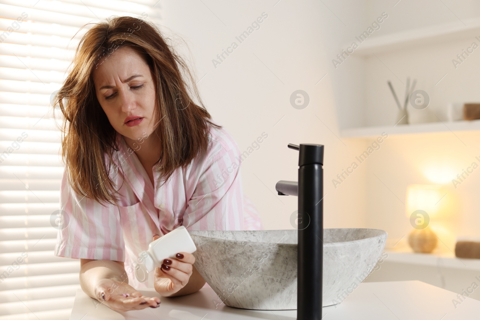 Photo of Unhappy woman with bottle of pills suffering from hangover near sink in bathroom