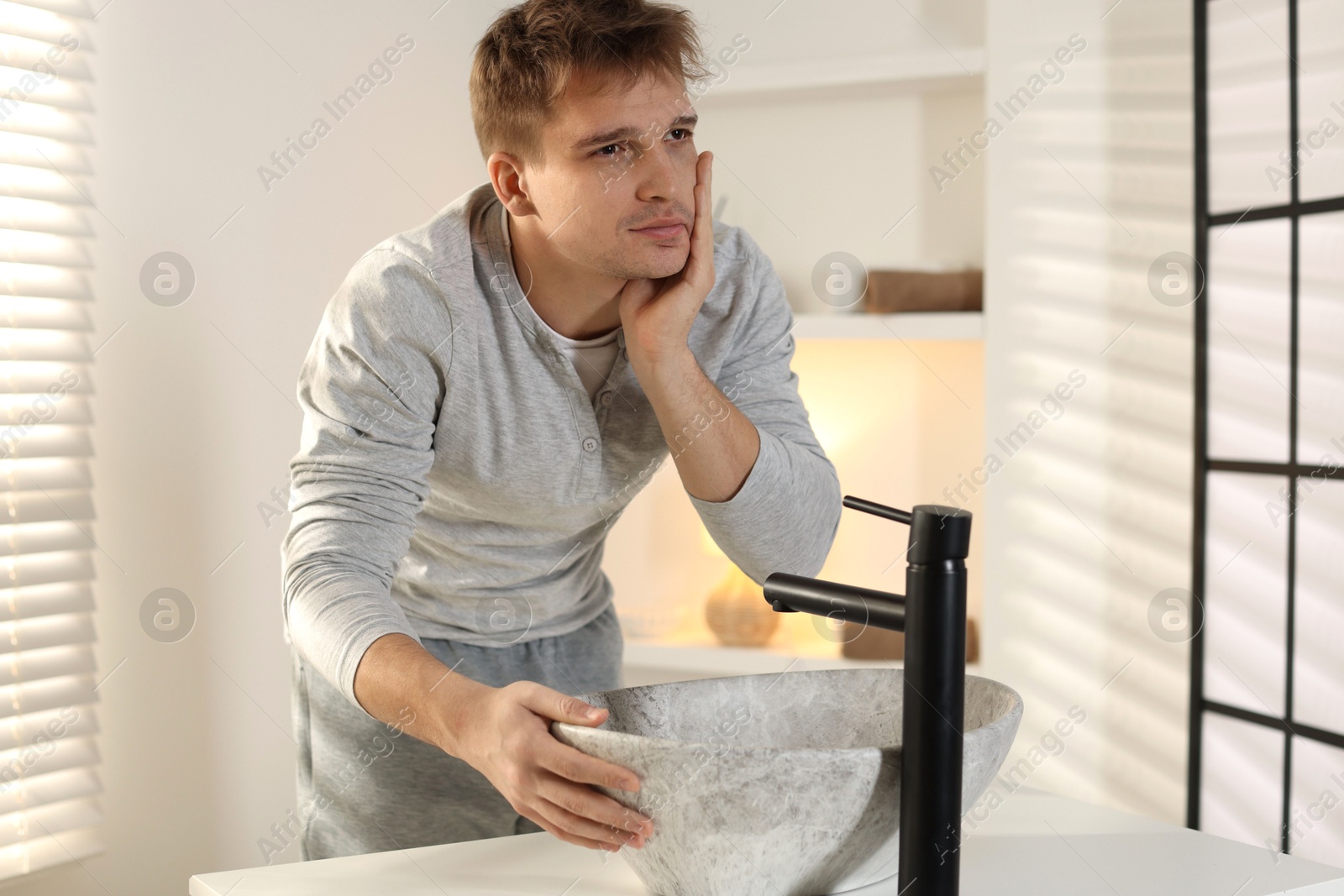 Photo of Unhappy young man suffering from hangover near sink in bathroom