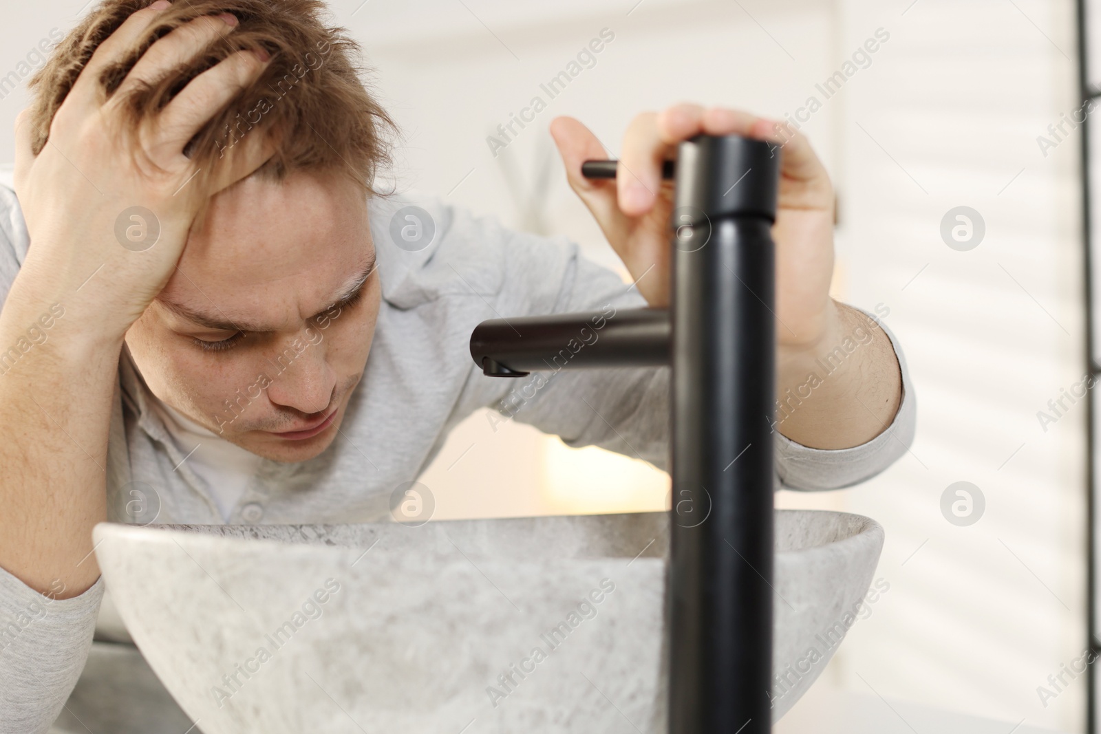 Photo of Unhappy young man suffering from hangover near sink in bathroom