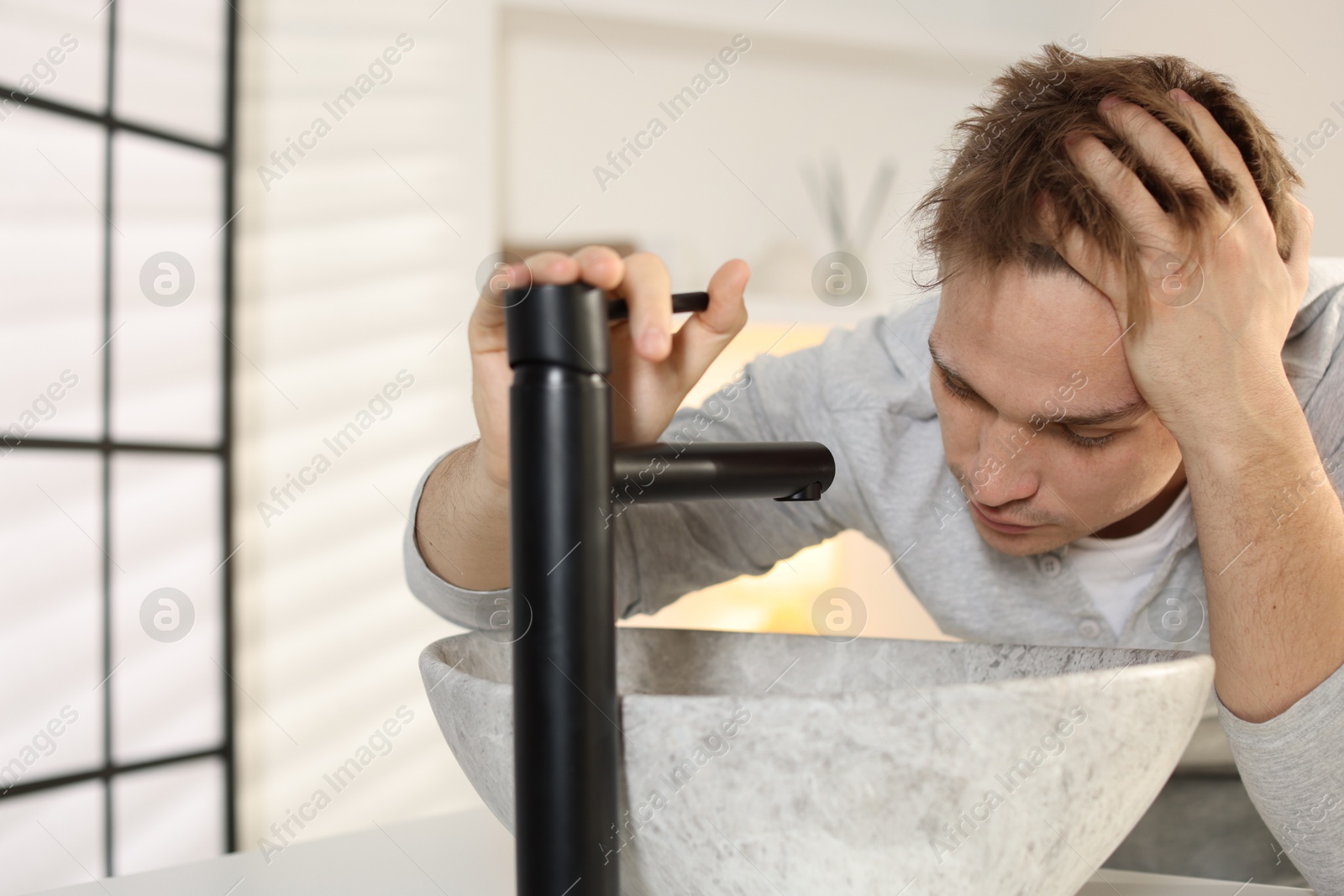 Photo of Unhappy young man suffering from hangover near sink in bathroom