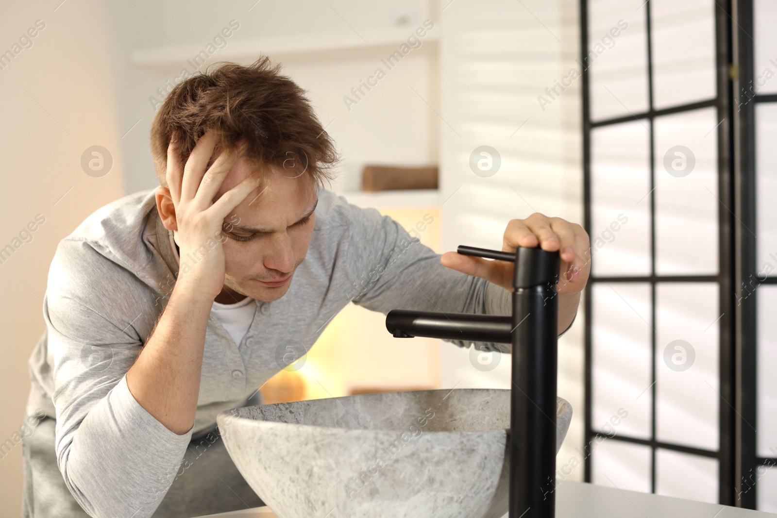 Photo of Unhappy young man suffering from hangover near sink in bathroom