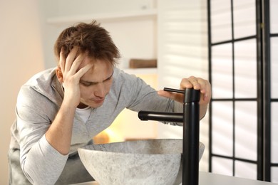 Unhappy young man suffering from hangover near sink in bathroom