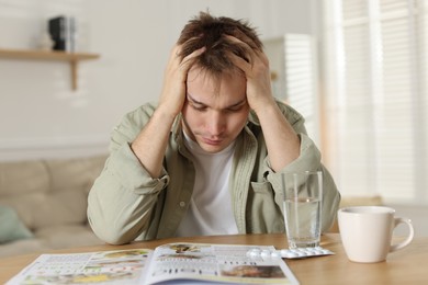 Suffering from hangover. Unhappy young man with cup of coffee sitting at wooden table indoors