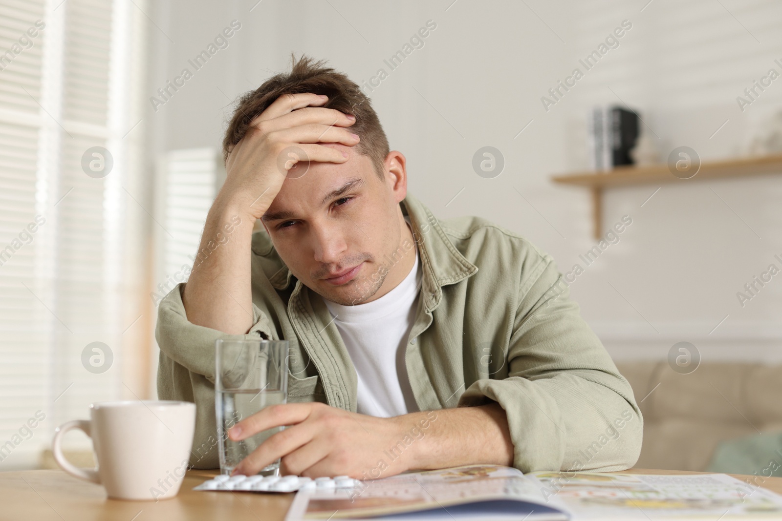 Photo of Suffering from hangover. Unhappy young man with cup of coffee sitting at wooden table indoors