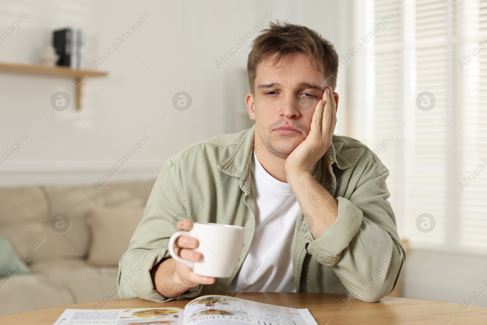 Photo of Suffering from hangover. Unhappy young man with cup of coffee sitting at wooden table indoors